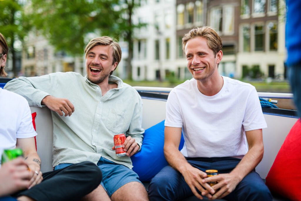 Twee personen die zich amuseren al zittend op een privé-boot Amsterdam met een biertje in hun hand. 